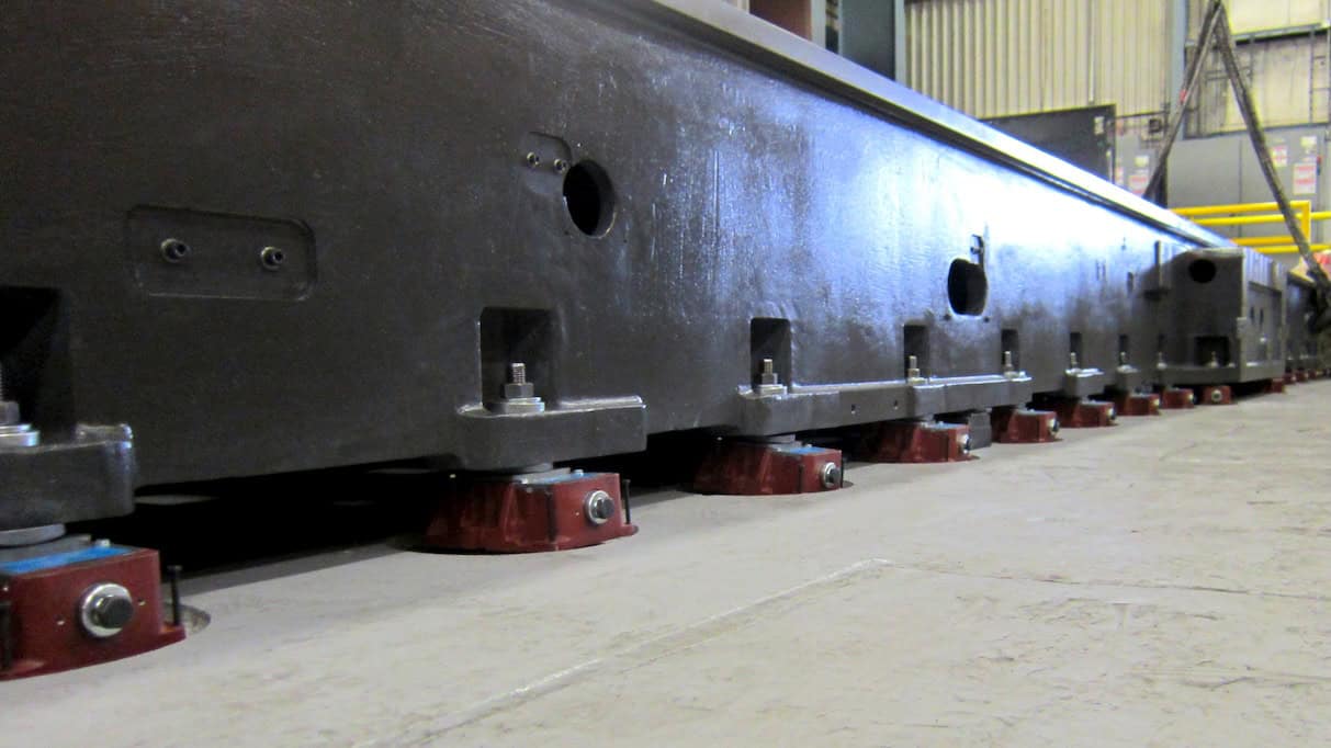 Close-up of a large, industrial metal structure mounted on red support feet, secured with bolts. The scene is indoors, with part of the warehouse visible in the background, including a ladder and yellow railing.