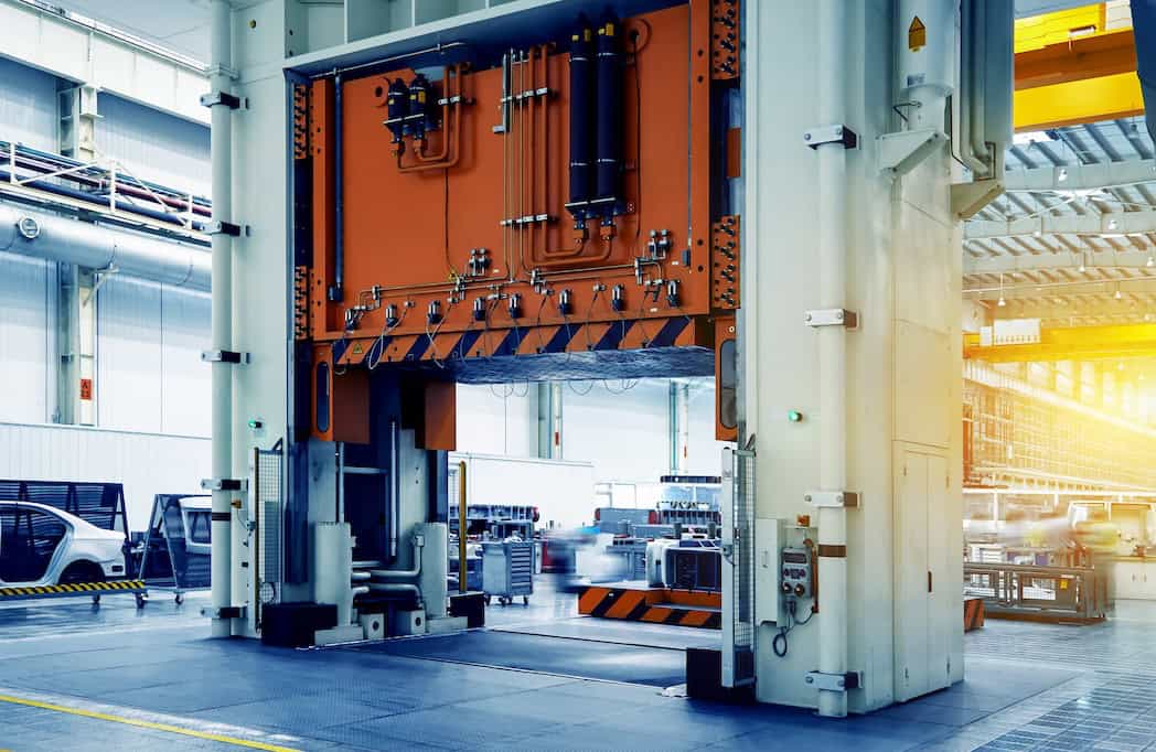 Large-scale punching lathes on automobile production lines A large industrial press machine in a factory setting, with a vibrant orange panel and hydraulic components. The background features a spacious area with a high ceiling, lit by natural and artificial light, and parked cars visible on the left.