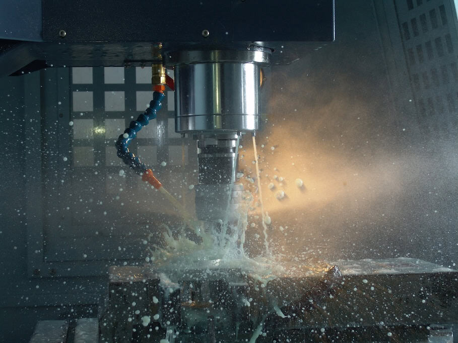 CNC Milling A close-up of a CNC machine in action, cutting metal. Coolant fluid is splashing around as the spinning tool works on the material, creating a dynamic display of motion and light reflection. The background is dark, emphasizing the machine's operation.