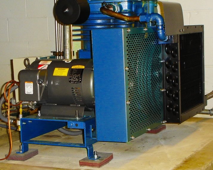 7-3-06 download 068 flipped A blue industrial air compressor with attached electric motor sits on a concrete floor in a utility room. The unit has ventilation grilles, cooling pipes, and is elevated on rubber pads.
