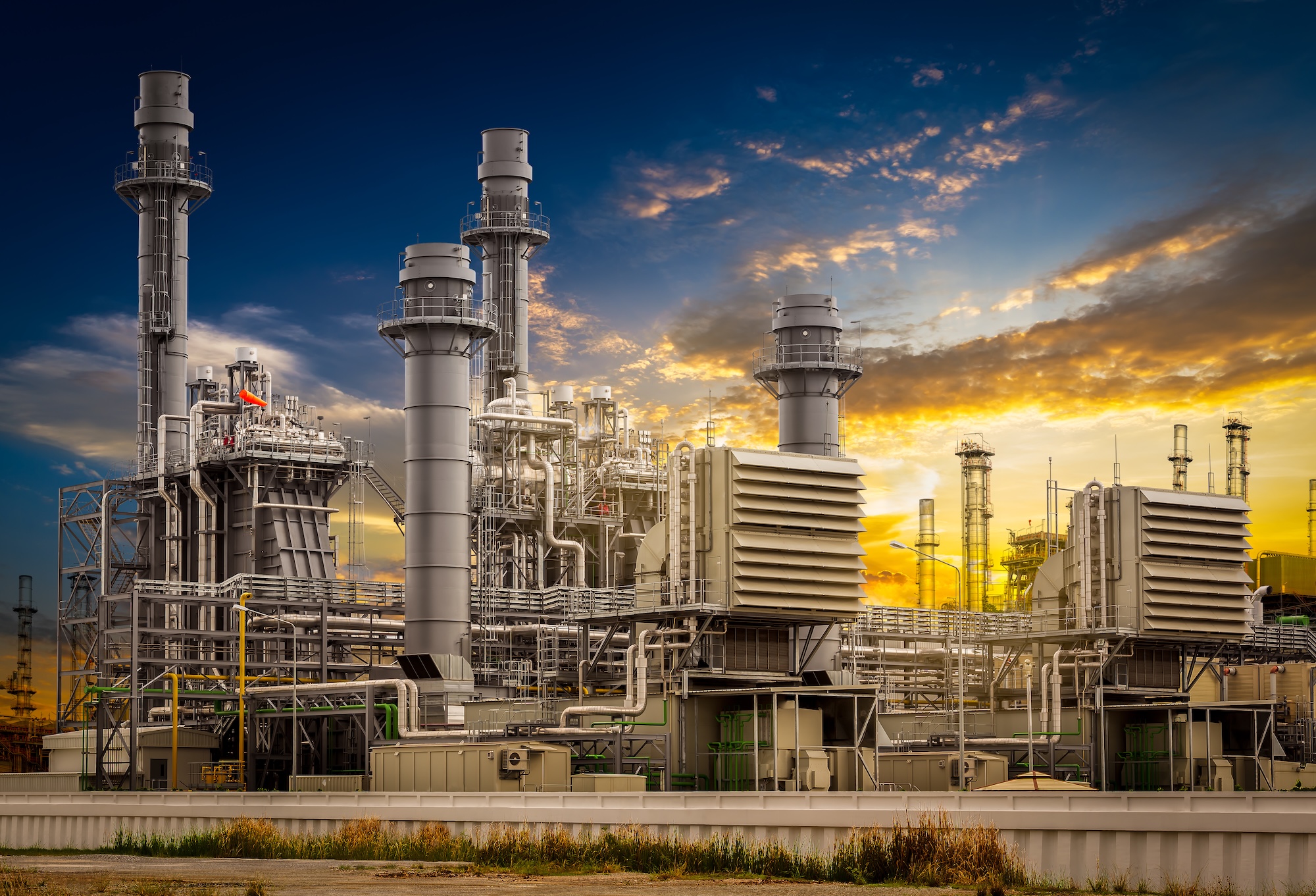 Power plant station A modern industrial power plant with large metal towers, pipes, and machinery, set against a dramatic sunset sky with clouds and warm light.