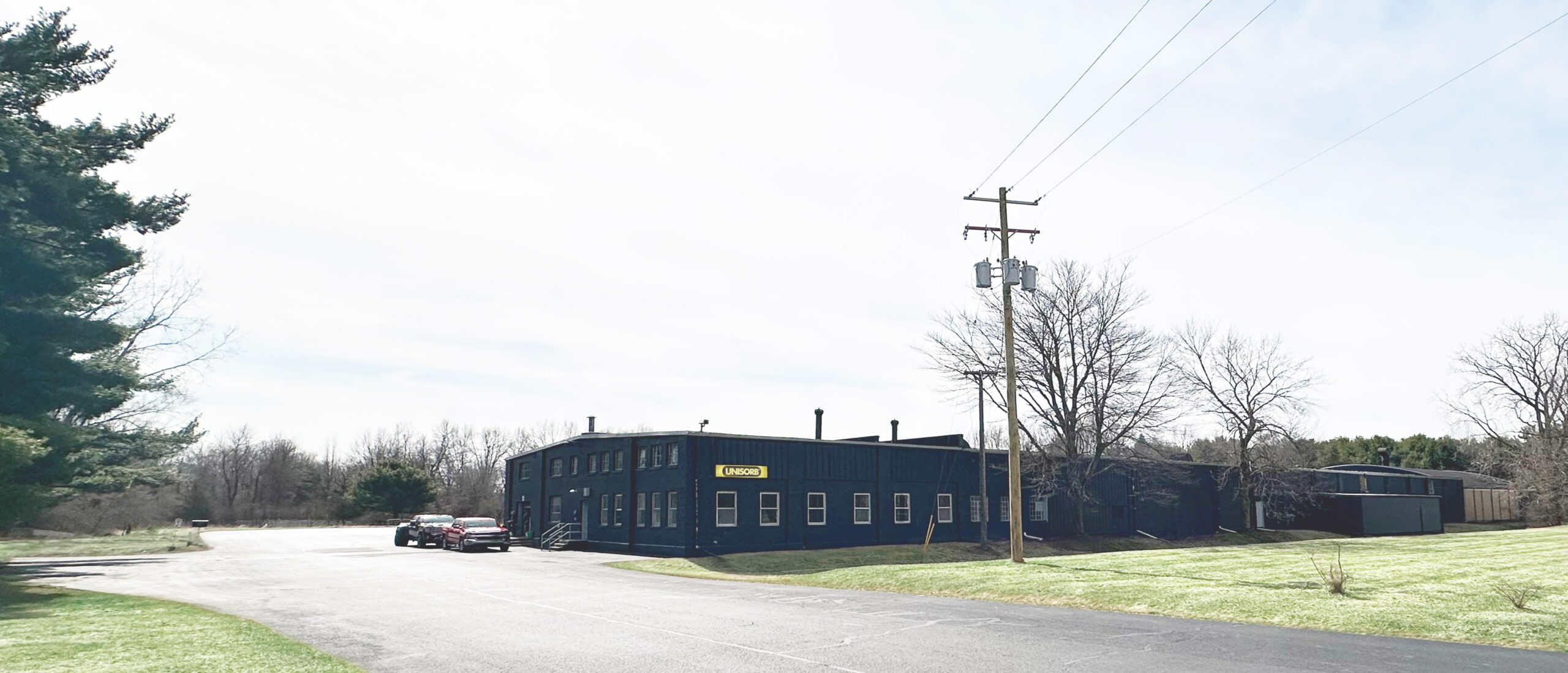 A dark blue, rectangular building with multiple windows sits near an empty parking lot with a few parked cars. Leafless trees and utility poles are nearby, and grassy areas surround the scene under a bright sky.