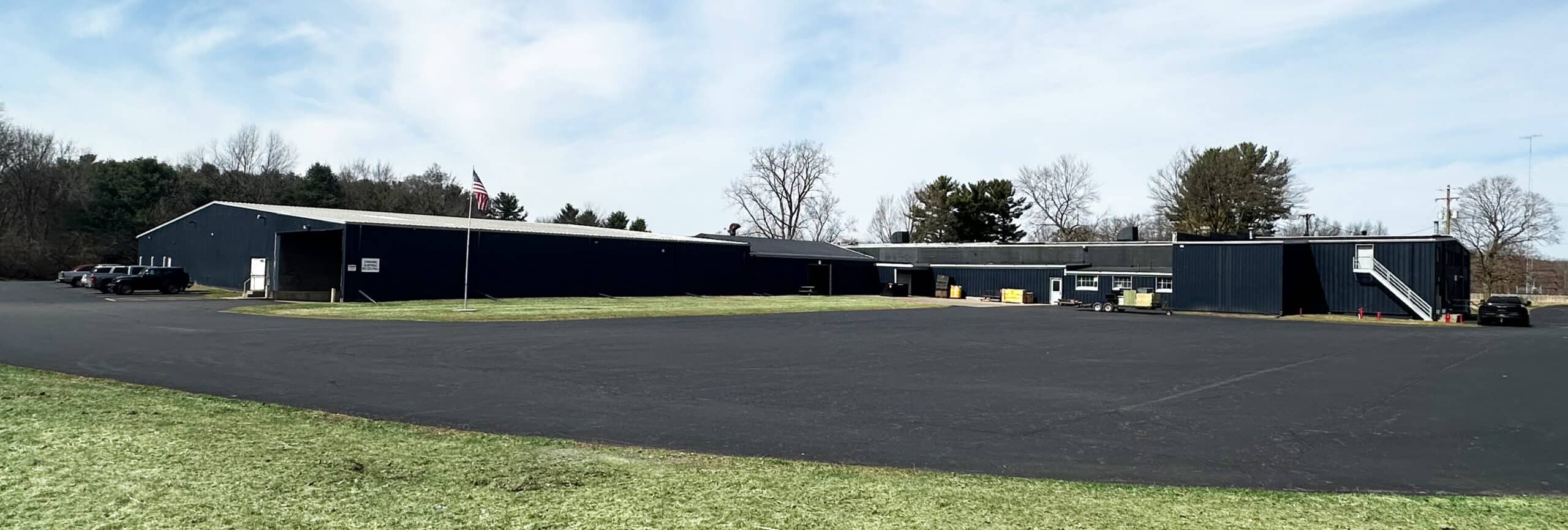 A large, single-story industrial building with dark exterior walls, surrounded by a wide, paved parking lot and bordered by grass and trees under a partly cloudy sky. Several vehicles are parked near the building.