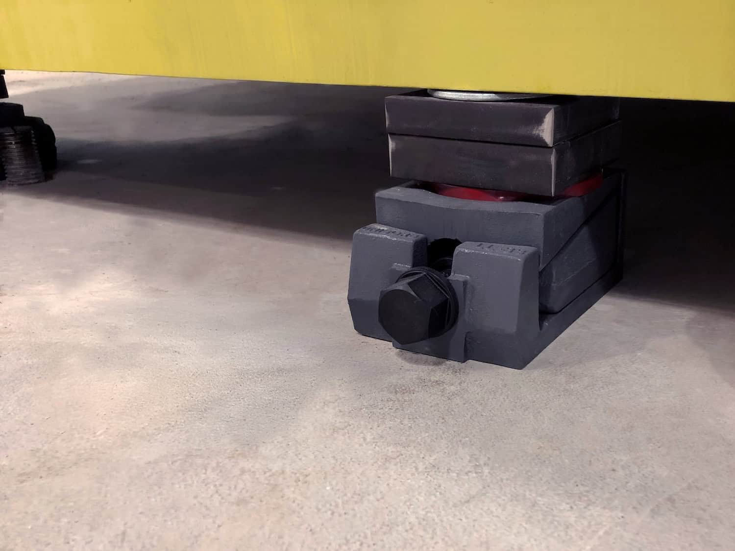Lev-l-ine Mount A large black leveling pad and a block are positioned under the corner of a yellow industrial machine, helping stabilize it on a concrete floor.