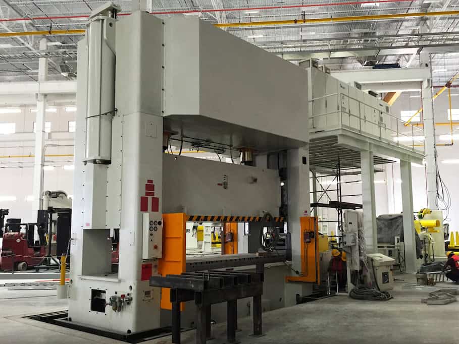 A large industrial press machine in a factory setting. The machine is gray with orange safety barriers. It occupies a spacious area with high ceilings and other machinery visible in the background.
