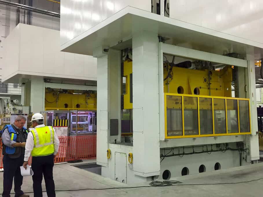 Two workers wearing safety vests and helmets stand in a large industrial facility. They are discussing near a massive, white and yellow machine. The area is surrounded by safety barriers and equipment.