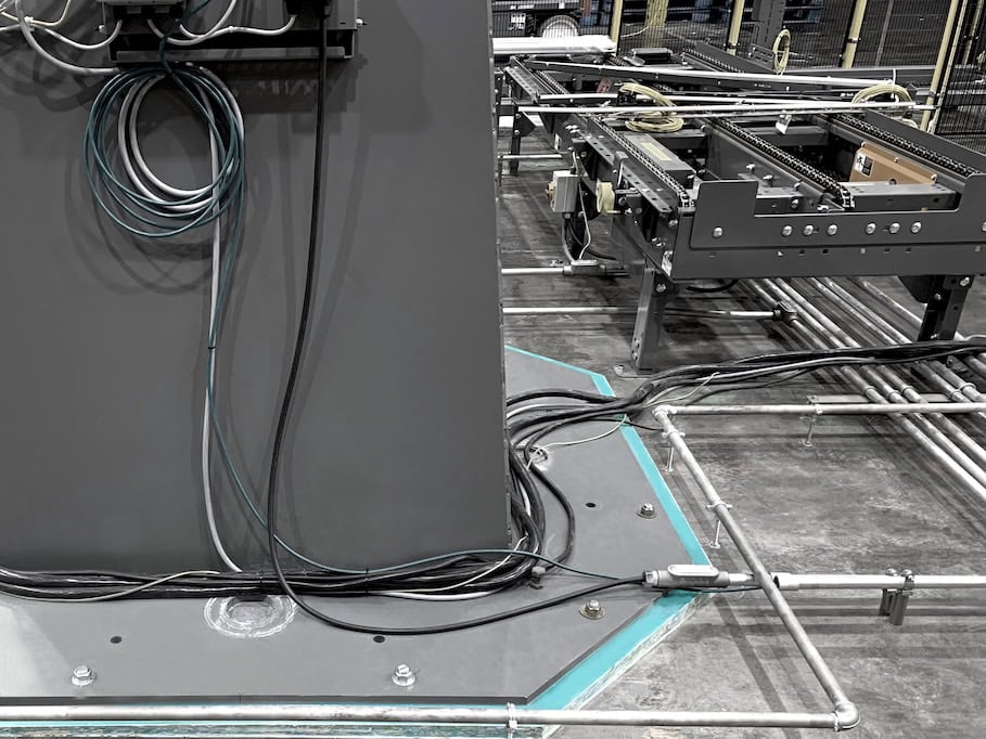 Grouting solutions. A complex network of cables is organized around a large industrial machine. Metal pipes and conduits run across the concrete floor. The machinery features various components and configurations, indicating a high-tech production environment.