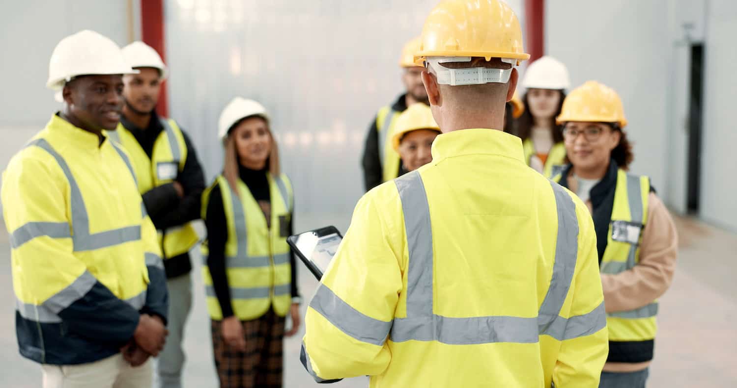Construction, teamwork and people in meeting for planning, brainstorming and discussion in building. Architecture, engineering and men and women for inspection, training development and project A group of construction workers in safety gear, including yellow reflective jackets and helmets, listen to a supervisor holding a clipboard during a meeting inside a building.