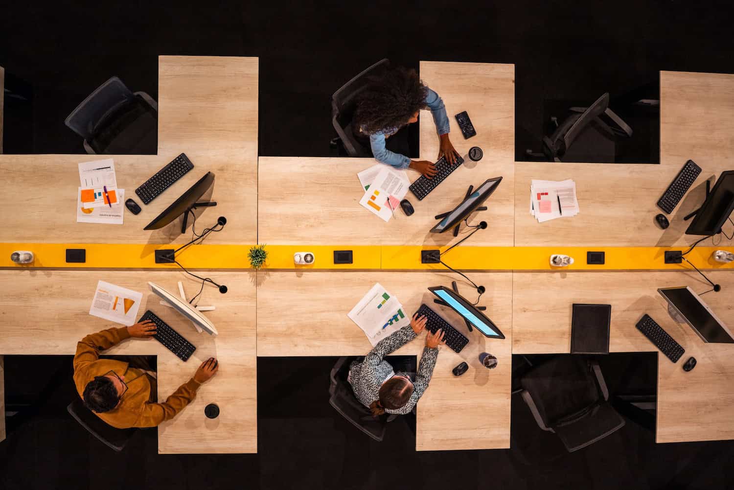 Coworkers talking and working using computer at office Overhead view of three people working at a shared office desk with computers, keyboards, documents, and coffee cups arranged neatly on a light wooden surface.