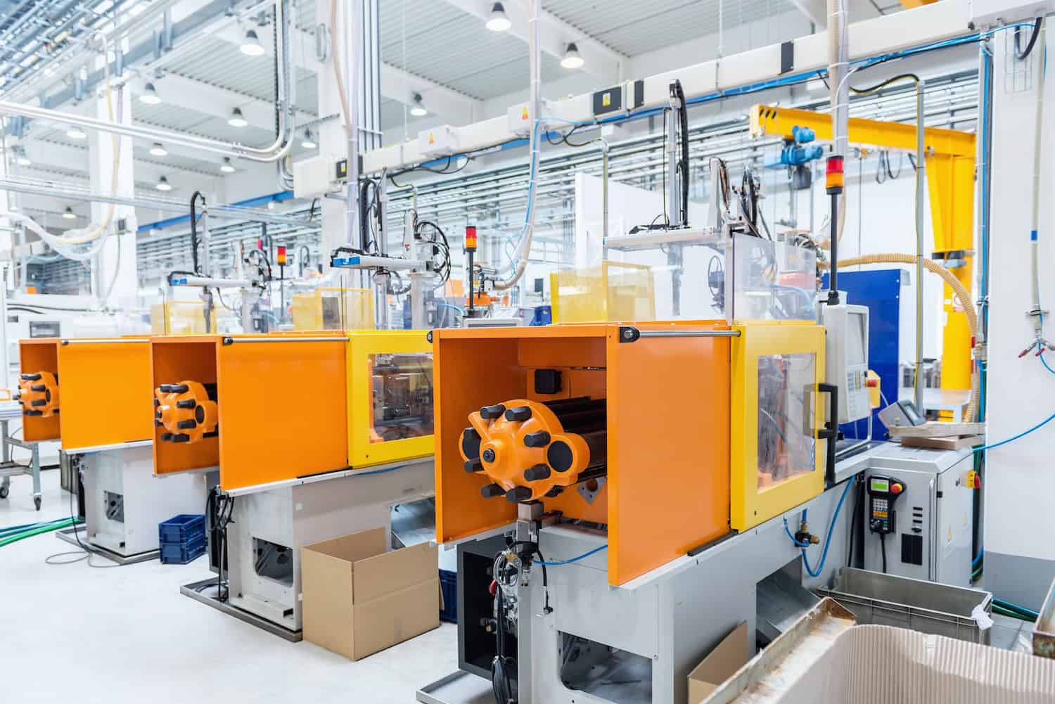 Futuristic injection moulding machines A row of modern orange and yellow industrial injection molding machines in a clean, brightly lit factory, surrounded by pipes, cables, control panels, and empty cardboard boxes.