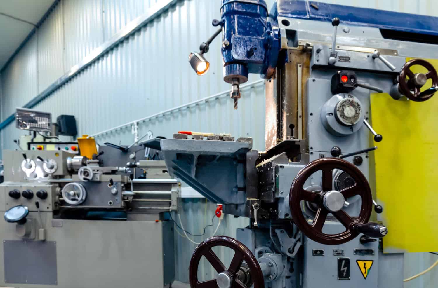 Shop for metal cutting A close-up view of industrial metalworking machines in a workshop, including a milling machine in the foreground and a lathe in the background, with blue walls and bright lighting.