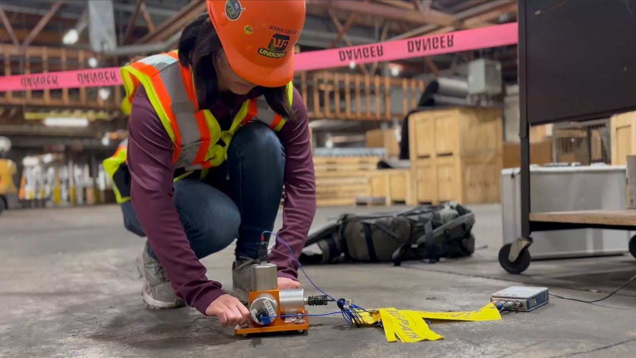 Vibration Survey A person wearing a hard hat and safety vest kneels on a warehouse floor, operating an orange device with attached wires and yellow tags. Crates, caution tape, and equipment are visible in the background.