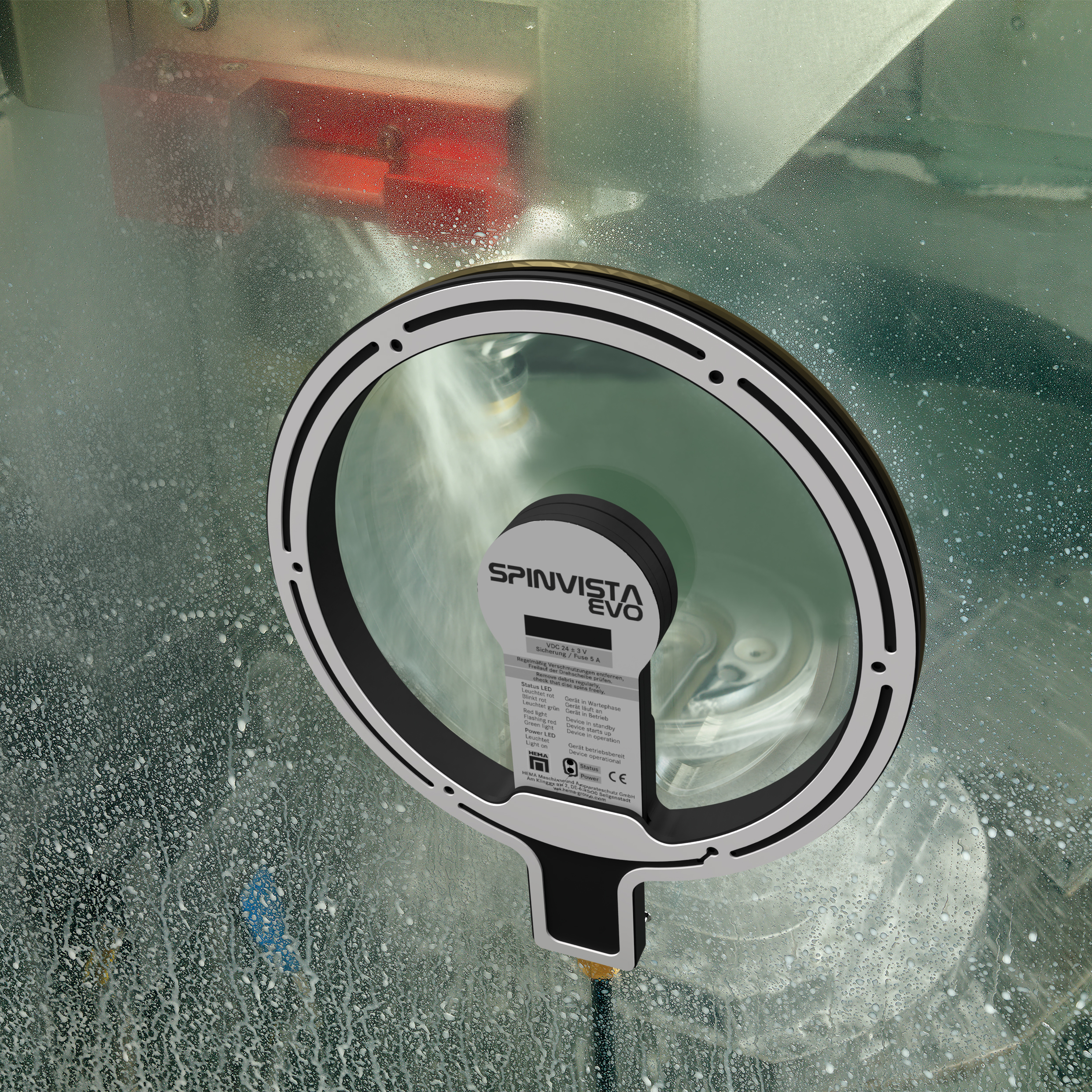 A round SpinView EVO inspection window with labels is mounted on a glass panel, showing a machine inside spraying water, with water droplets covering the glass surface.