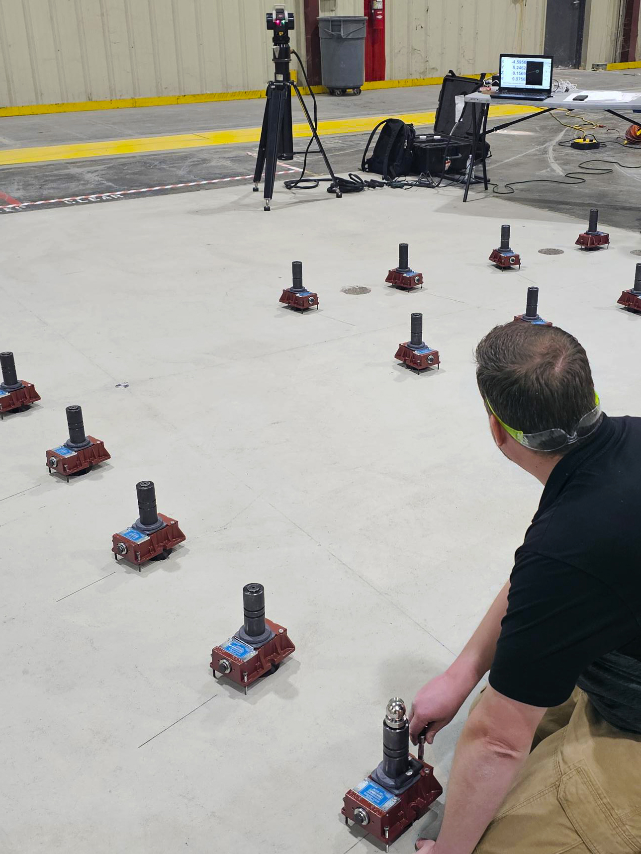 A man crouches on a large indoor concrete floor, adjusting one of several red RK Fixators arranged in a grid. A tripod with equipment and a laptop are set up nearby, with a yellow line and beige wall in the background.