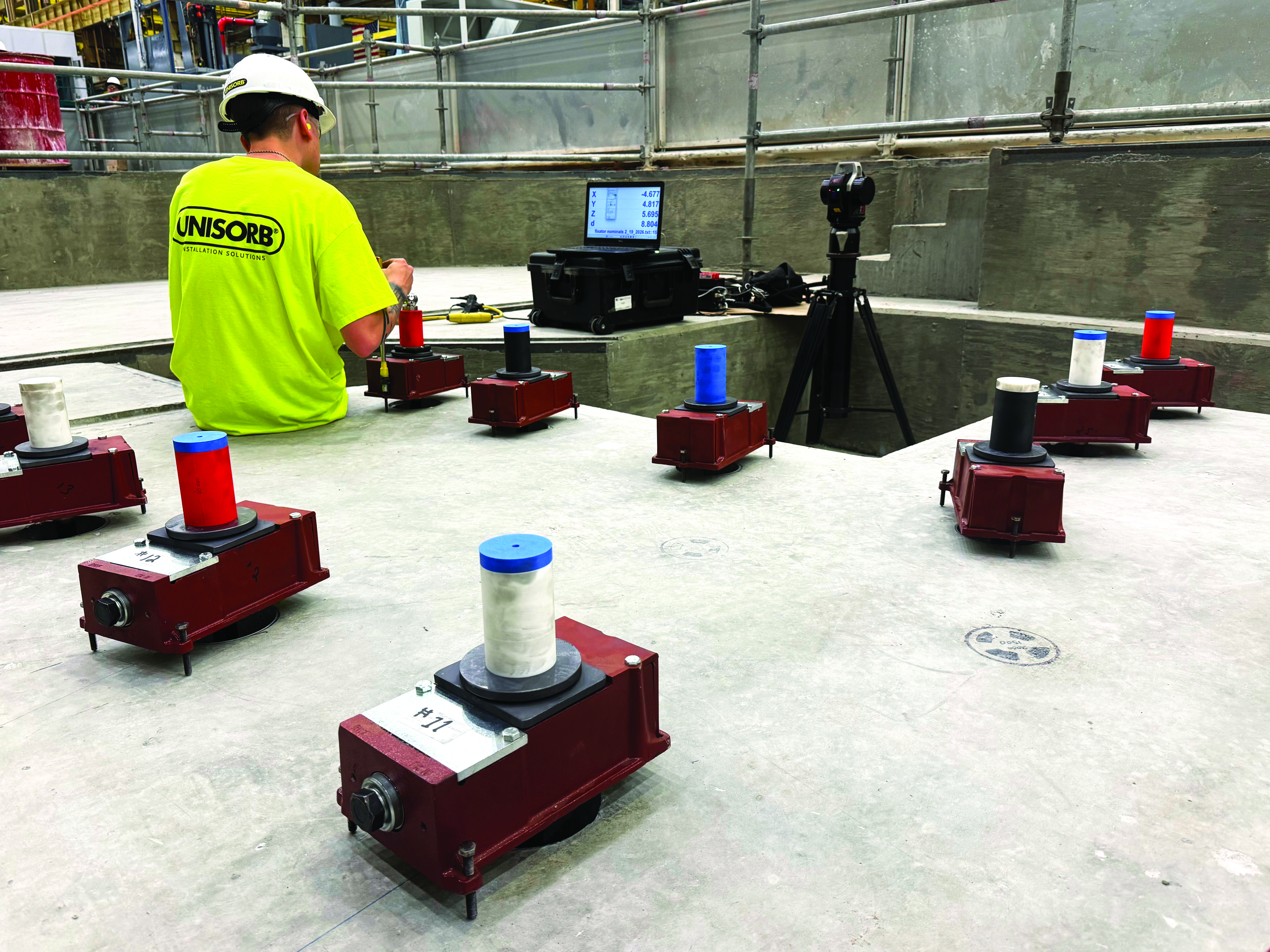 Multiple industrial machines or mechanical devices, each placed inside a white plastic container, are arranged in evenly spaced rows on a concrete floor in a large indoor facility.
