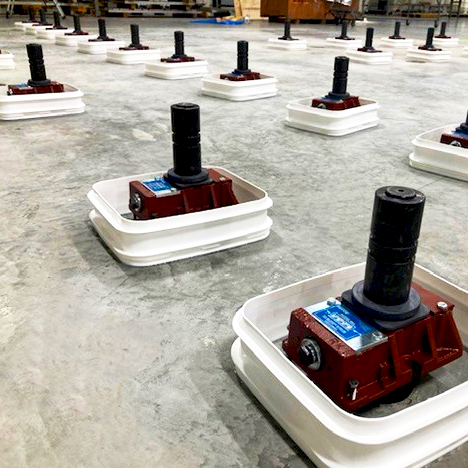 Multiple industrial machines or mechanical devices, each placed inside a white plastic container, are arranged in evenly spaced rows on a concrete floor in a large indoor facility.