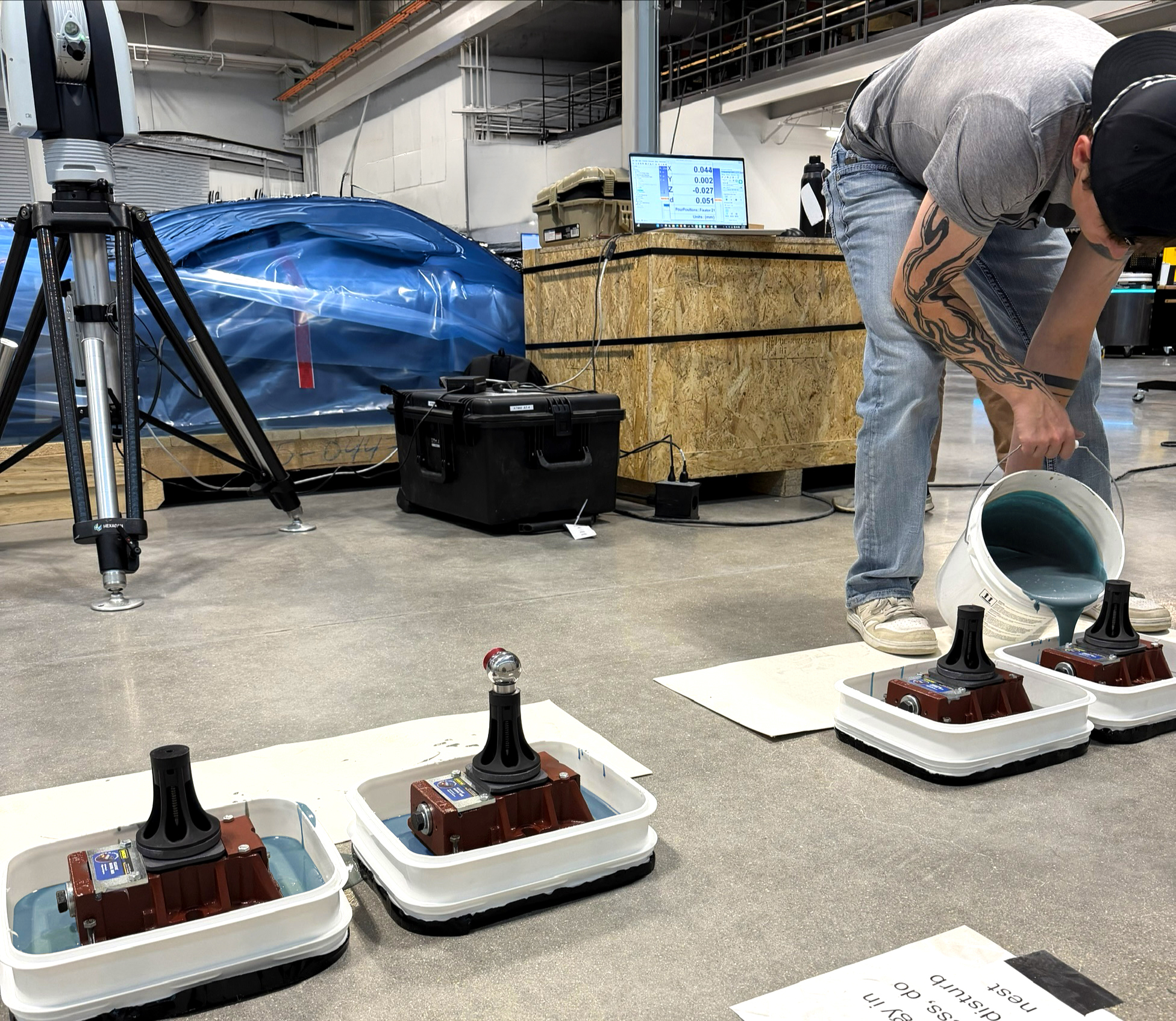 A person pours blue liquid from a bucket into containers holding mechanical sensor bases on a workshop floor.