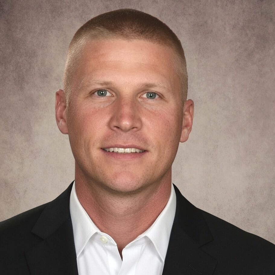 A man with short blonde hair, wearing a white dress shirt and a black suit jacket, poses in front of a mottled gray background, smiling slightly at the camera.